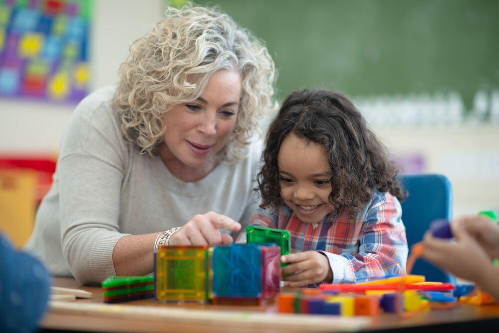 Woman leaning over table with a small smiling girl sitting at it; the girl is playing with brightly coloured building