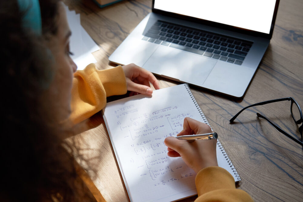 Woman at a wooden desk writing in a notepad with a laptop also on the table