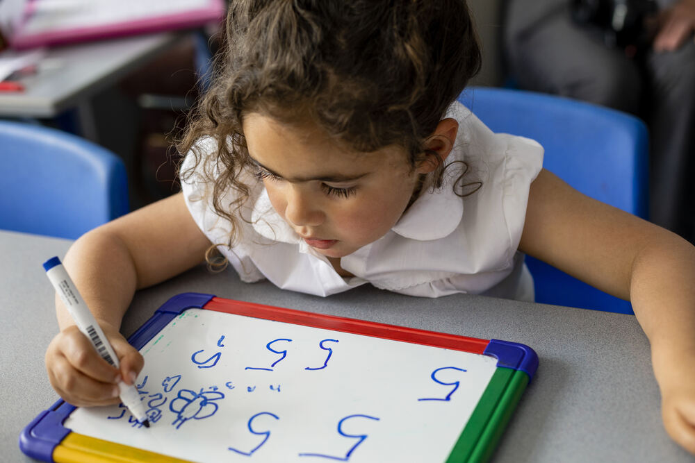 Primary school girl sitting at a table, writing numbers on a white plastic drawing tablet