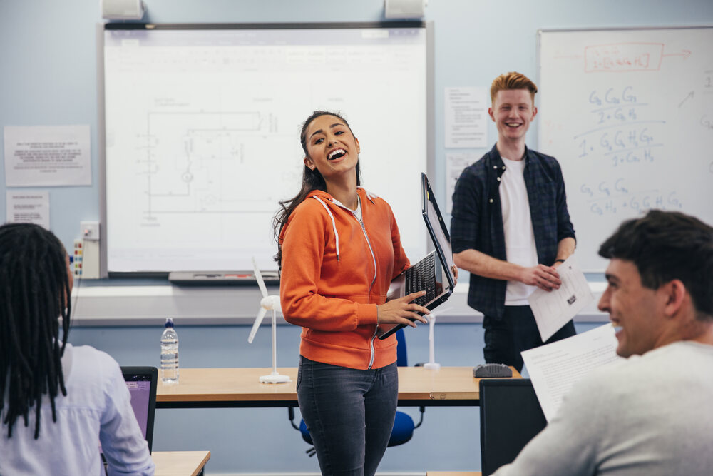 Woman at the front of the class wearing bright orange and laughing, holding a laptop, with three other people also in the photo